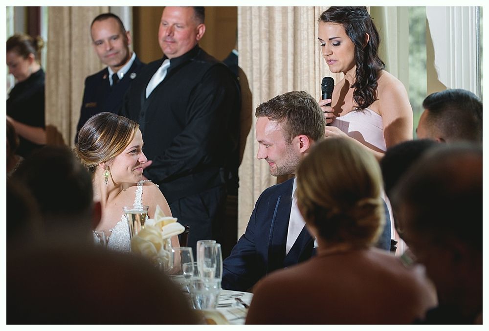 Bride and groom at a wedding, listening to a speech. A woman speaks into a microphone.