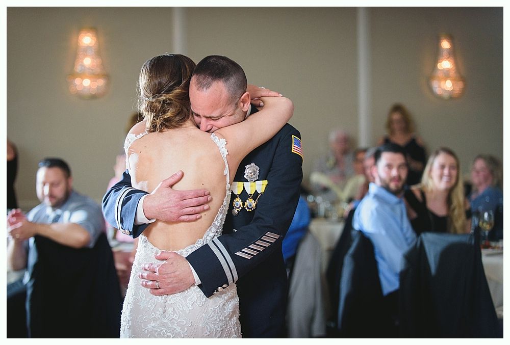 Bride and military officer embracing and dancing at a wedding reception; guests watch.