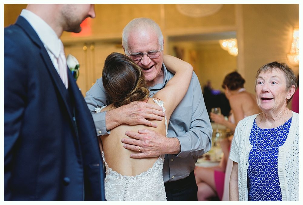 Bride hugs a man, likely her father, at a wedding reception. Woman watches with a surprised expression.