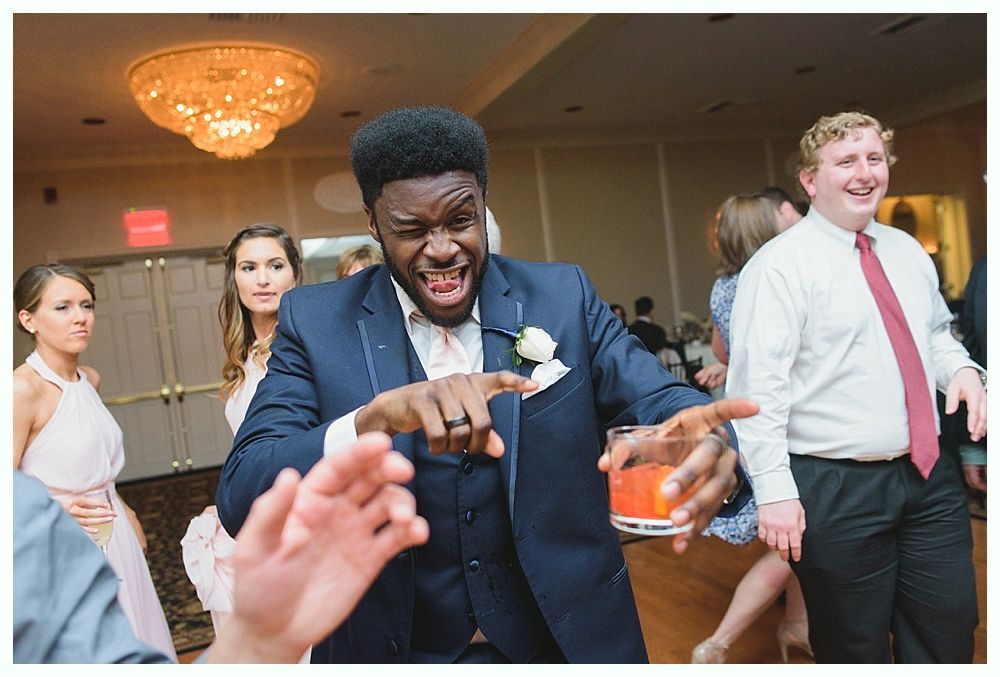 Man in a suit dancing with a drink, pointing. Others in formal wear at a wedding reception.