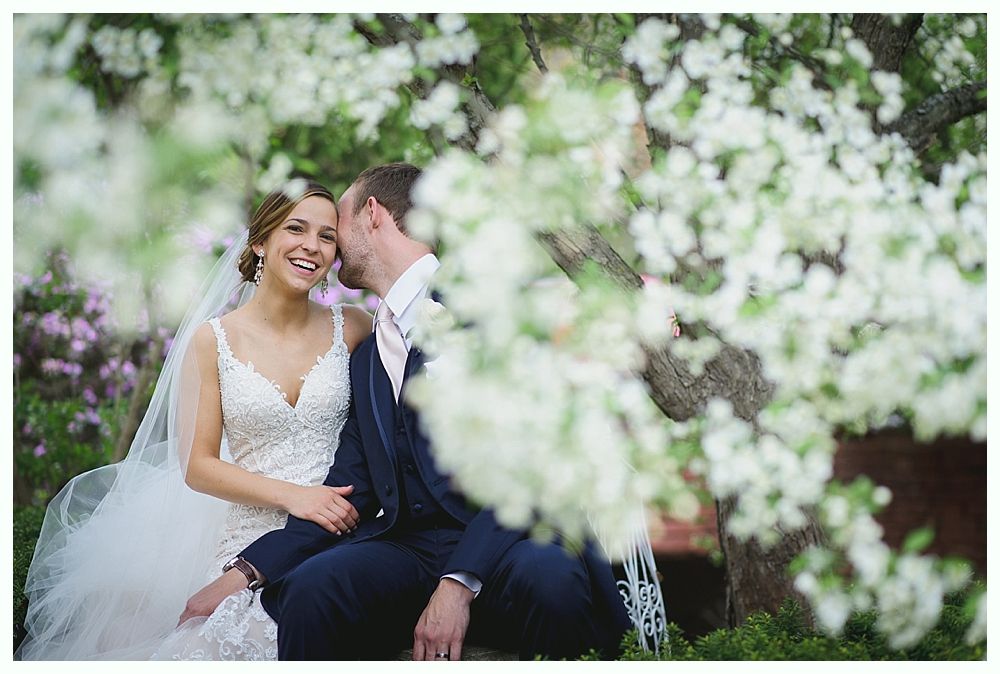 Bride and groom seated outdoors, surrounded by white flowers. Groom whispers to bride, who smiles.