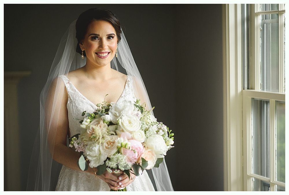Bride in a white lace wedding dress, veil, and holding bouquet, smiles near window.