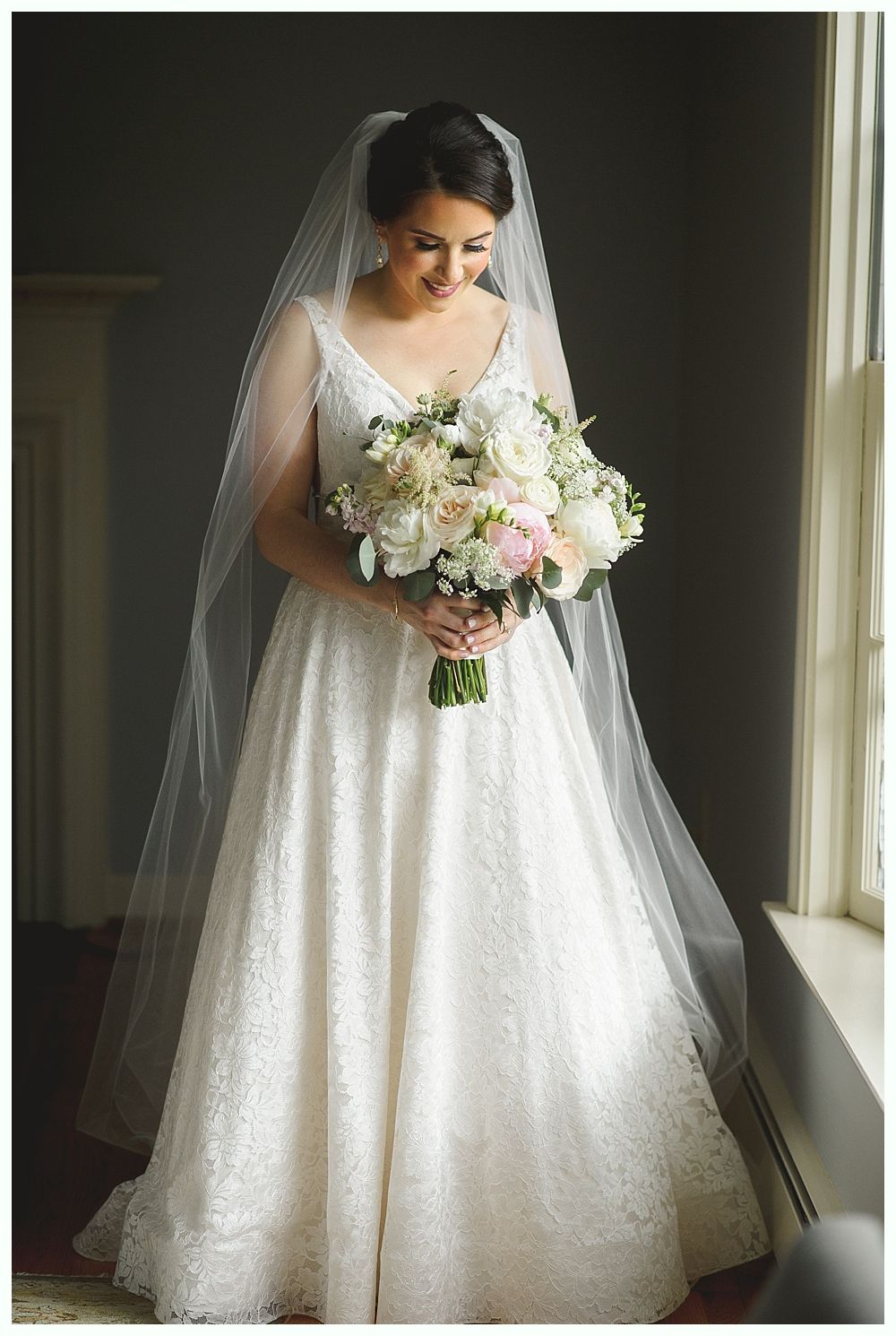Bride in white lace wedding dress holding bouquet, smiling, by window.