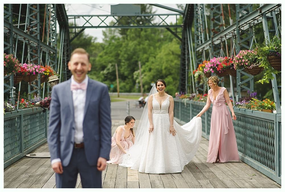 Groom smiles at camera on a bridge. Bride and bridesmaids pose, adjusting the wedding dress. Colorful flowers decorate the bridge.