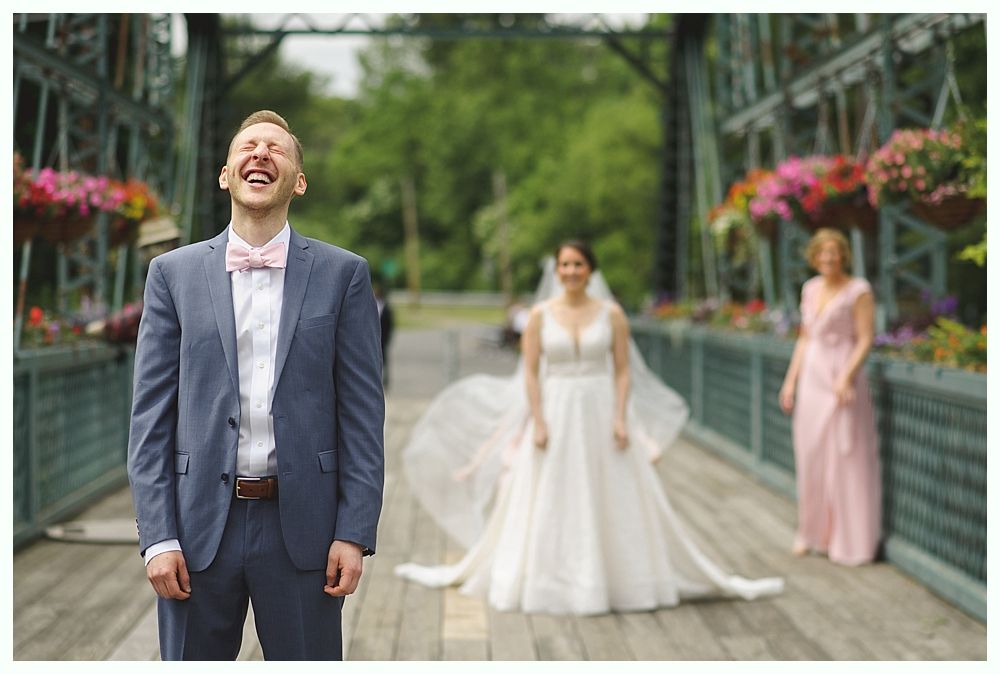 Groom laughs, bride and woman in pink stand on a bridge decorated with flowers.