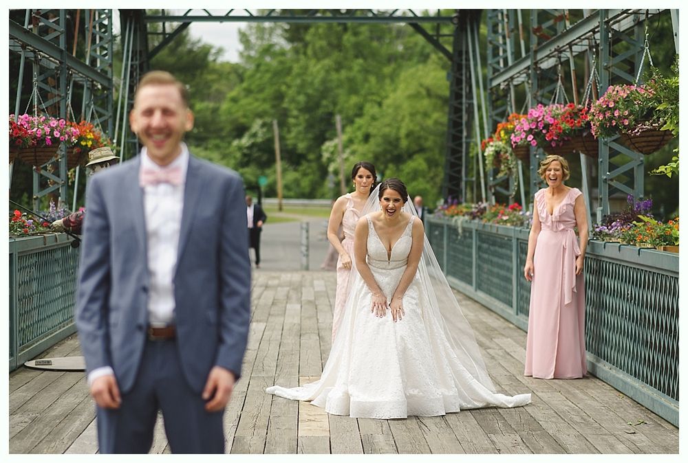 Groom smiles at bride in white dress on bridge, bridesmaids and flowers nearby.