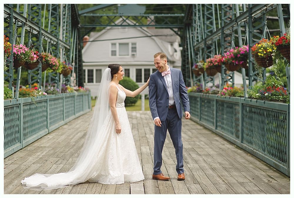 Bride and groom on a bridge. She touches his face, laughing. Surrounded by hanging flower baskets.