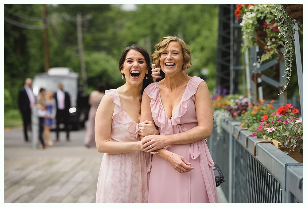 Two women in pink dresses laughing on a bridge with flowers; wedding party in the background.