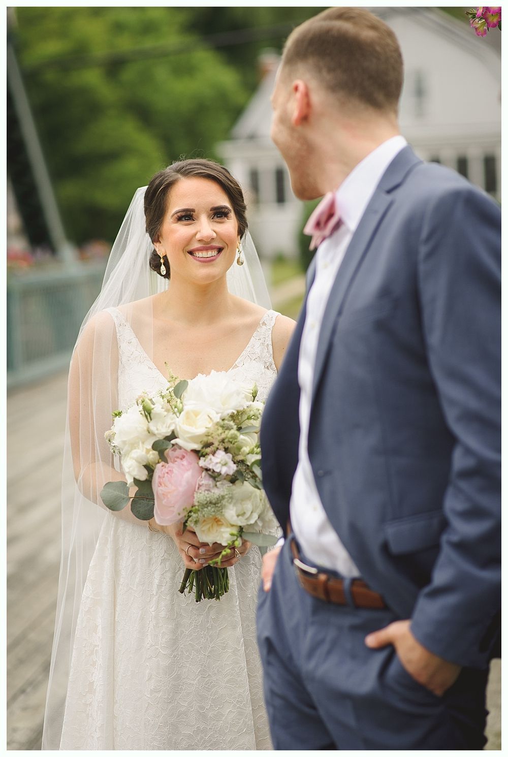 Bride smiles at groom, holding a bouquet. She wears a white dress and veil. He wears a blue suit, looking at her.