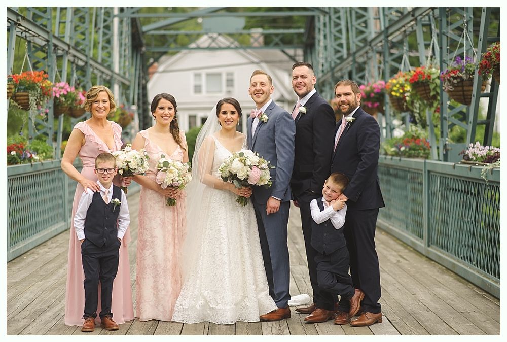 Wedding party on a bridge, smiling. Bride and groom centered, surrounded by bridesmaids and groomsmen, children.