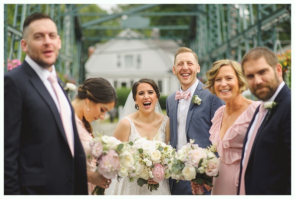 Wedding party laughs on a bridge. Bride holds flowers, wearing a veil. Groomsmen in suits. Bridesmaids in pink.