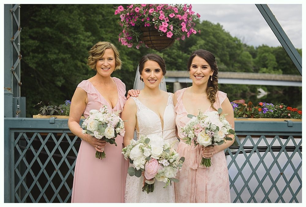 Bride and bridesmaids pose on a bridge with flower bouquets. They wear pink dresses.