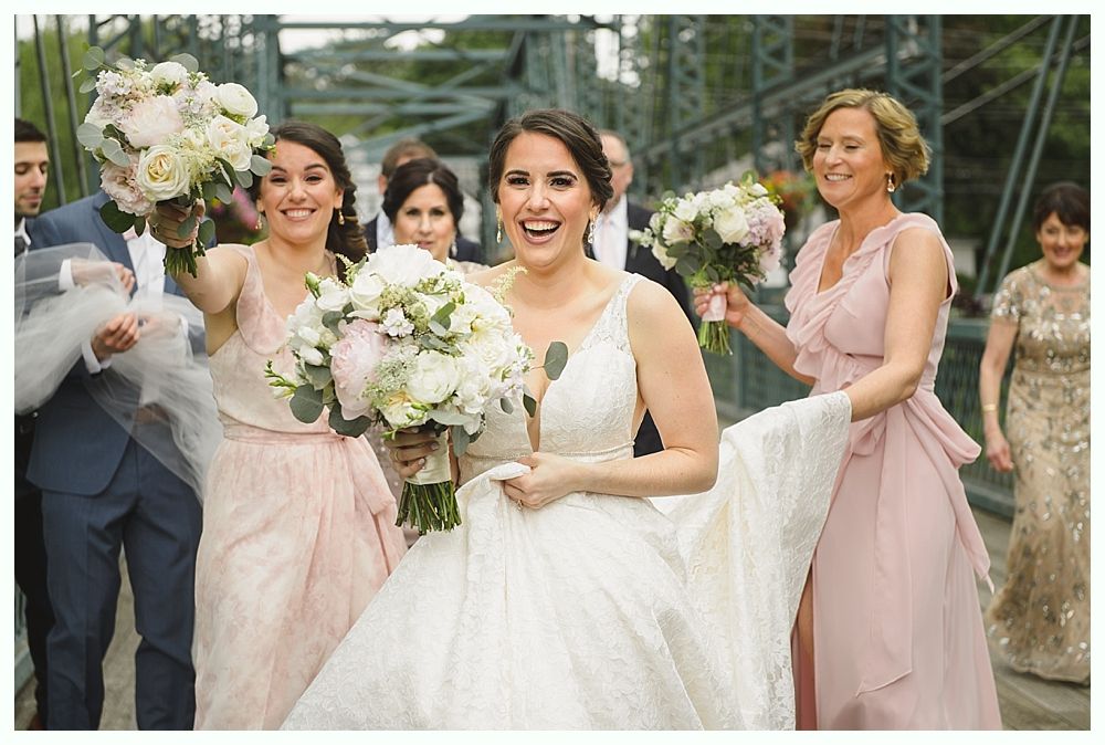 Bride in white dress on bridge, smiling with bridesmaids holding bouquets.