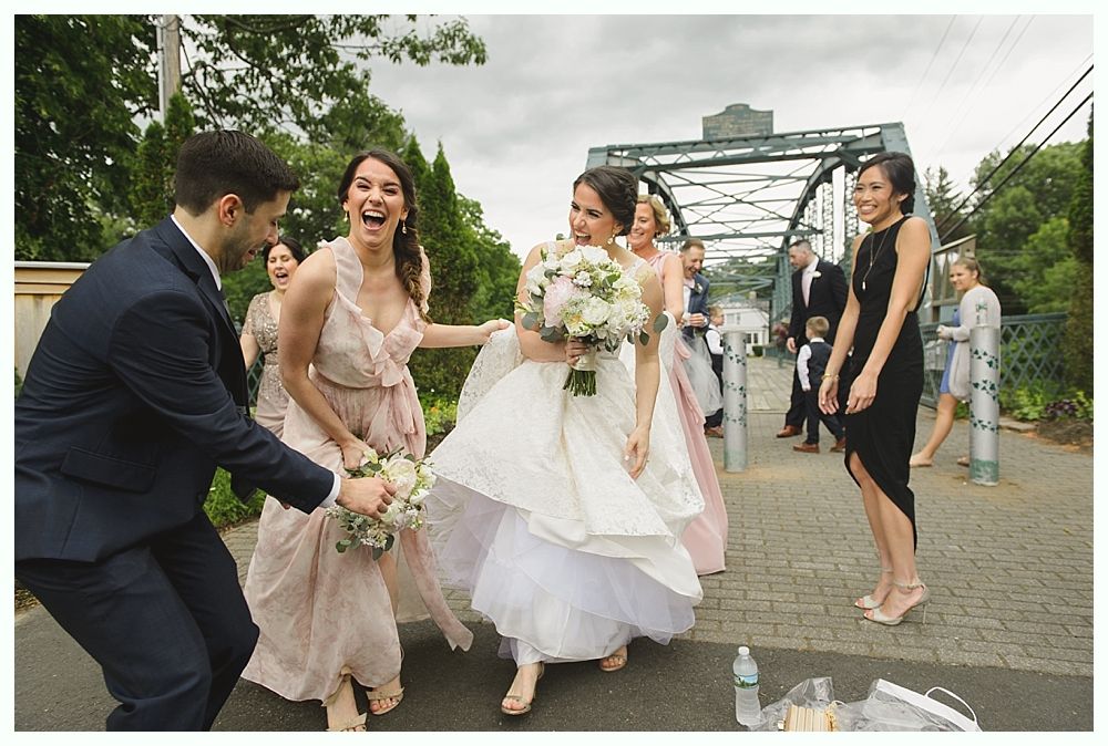 Wedding party laughing and celebrating on a bridge; bride holding bouquet, groom dancing, bridesmaids smiling.
