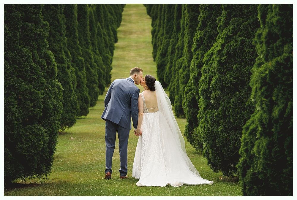 Couple kissing, walking down a path flanked by tall green trees; bride in white gown, groom in blue suit.
