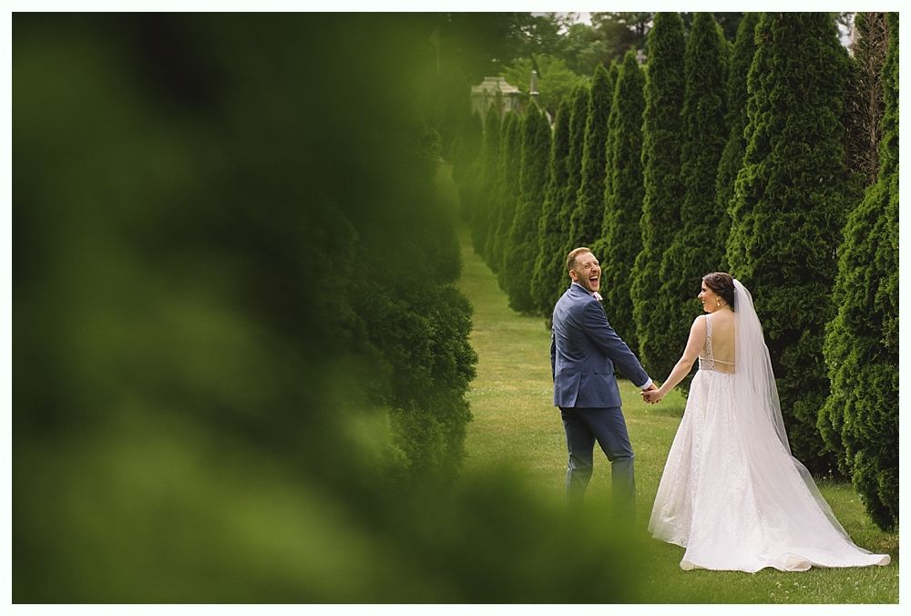 Bride and groom holding hands, walking in a park with rows of tall green trees; groom makes a surprised expression.