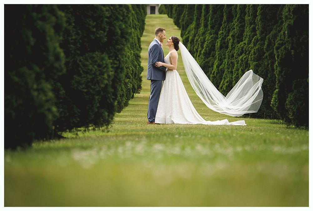 Couple in wedding attire embrace on a grassy path lined with green trees; the bride's veil flows in the air.