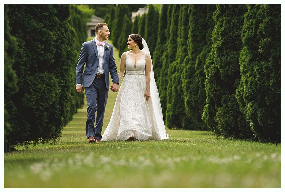 Couple in wedding attire walk hand-in-hand down a grassy path lined with tall, green trees, smiling at each other.