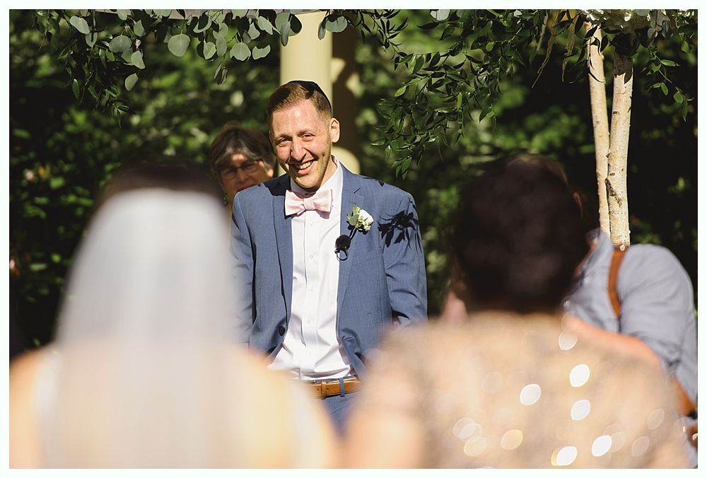 Groom smiles during wedding ceremony outdoors. He wears a blue suit, pink bow tie, and white shirt.