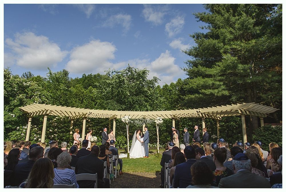 Outdoor wedding ceremony under a wooden arbor on a sunny day. Guests seated, couple at altar.
