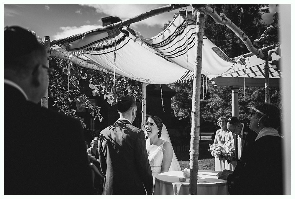Bride and groom under a chuppah during a Jewish wedding ceremony, smiling, outdoors.