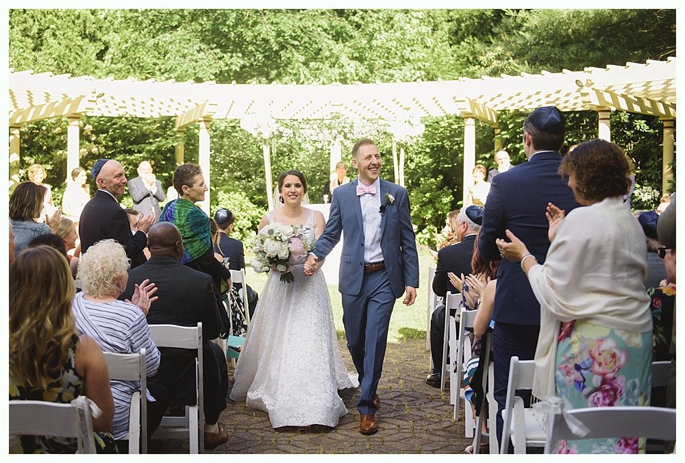 Newlyweds walking down the aisle, holding hands, cheered by guests. Outdoor wedding.