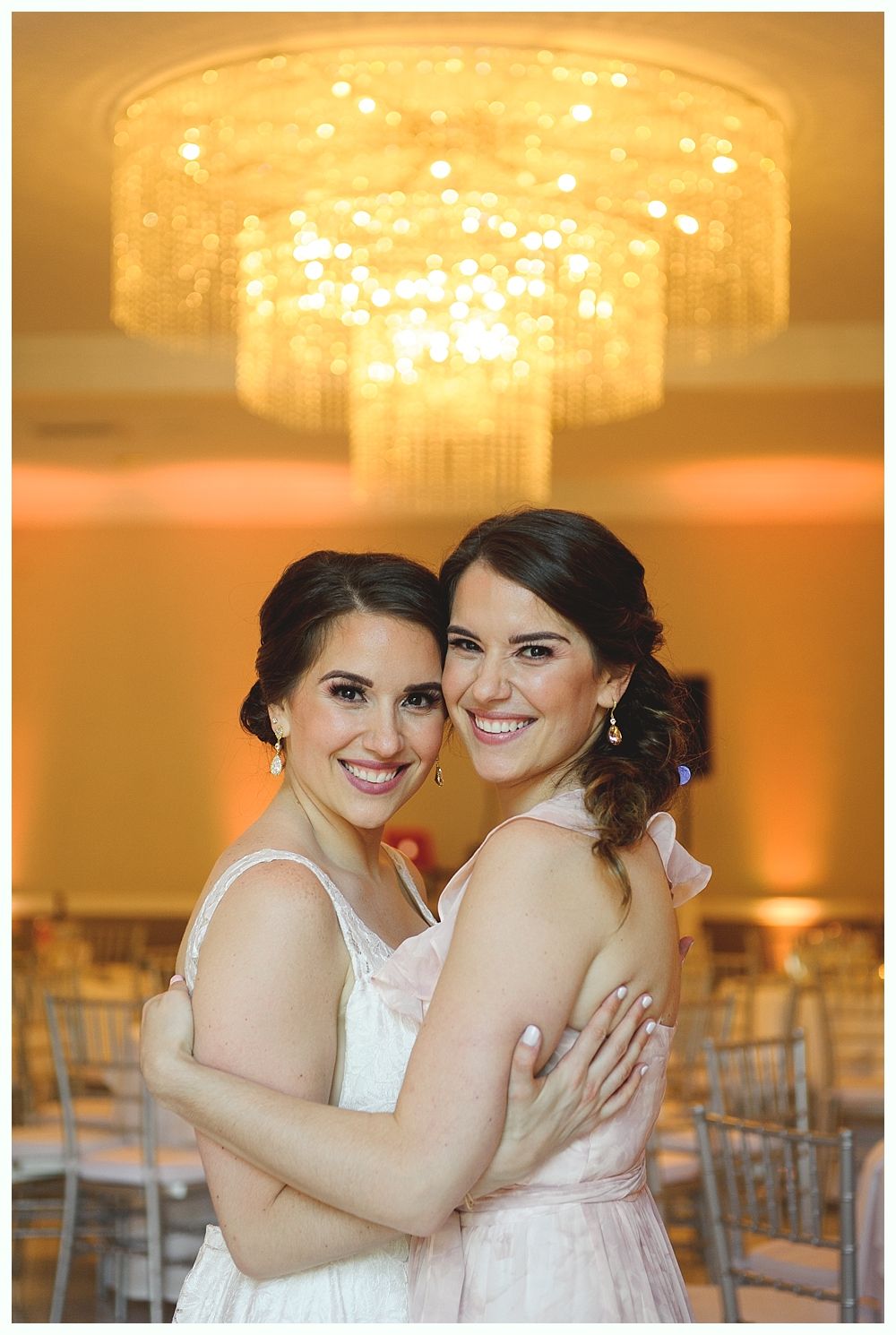 Two women in wedding attire embrace under a large chandelier; smiles.