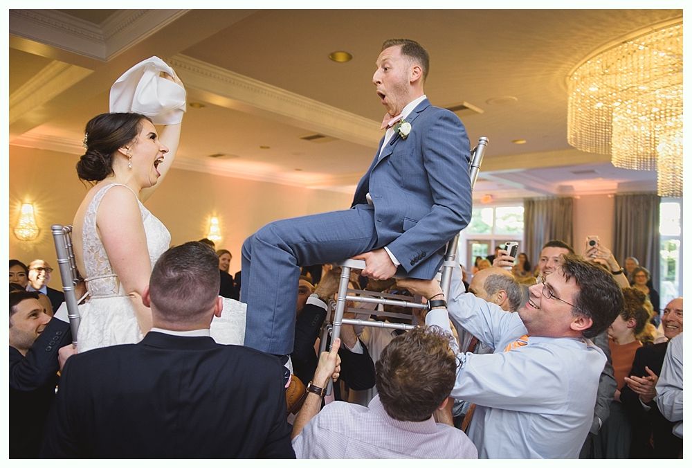 Wedding guests lifting bride and groom on chairs, celebrating indoors.