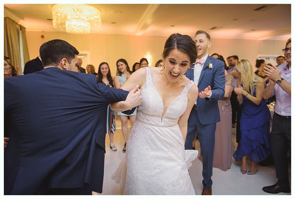 Bride being pulled by person in suit on dance floor, guests clapping and celebrating in ballroom.