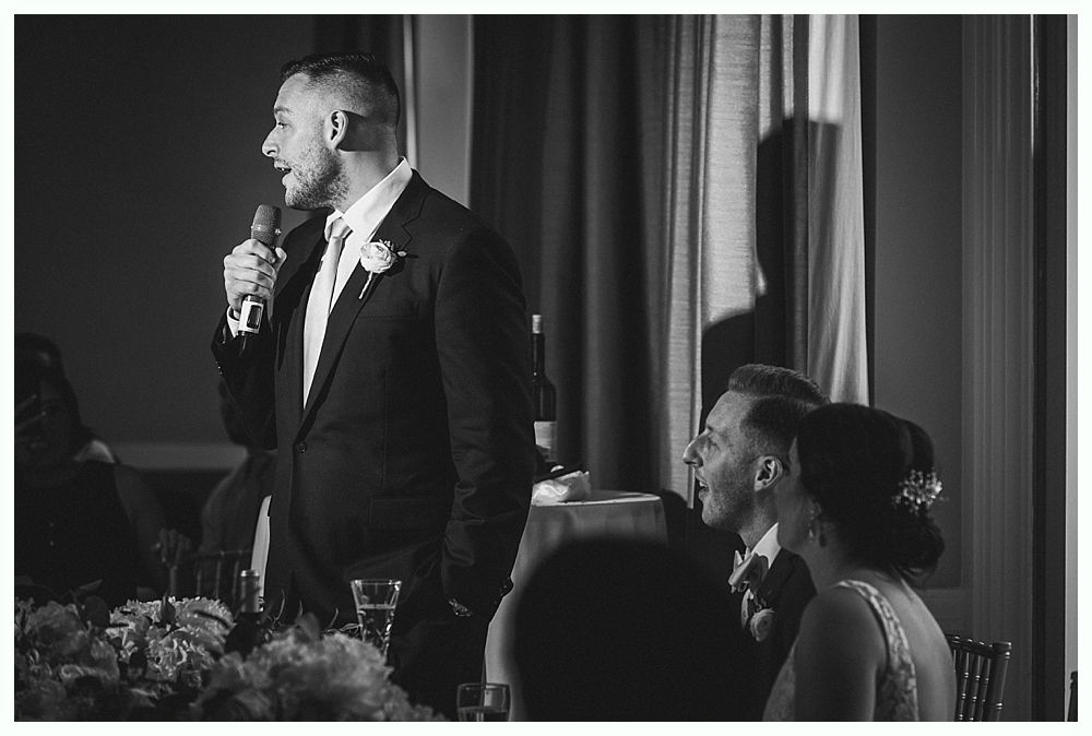 Man in suit gives a speech with a microphone; bride and groom listen at a table in a room.