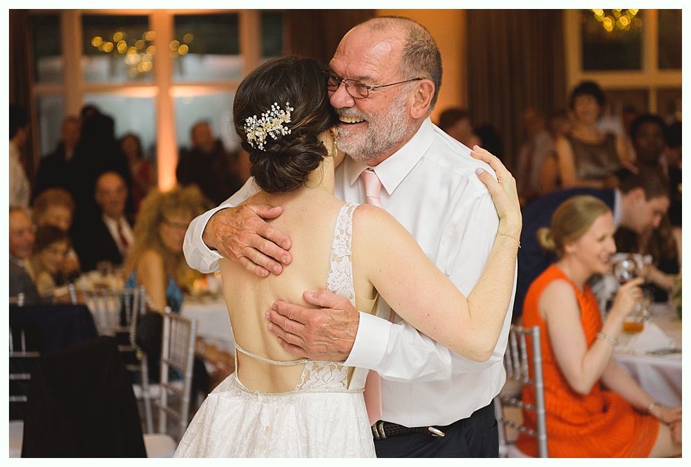 Bride in white dress dancing with older man, embracing, smiles. Reception setting, guests in background.