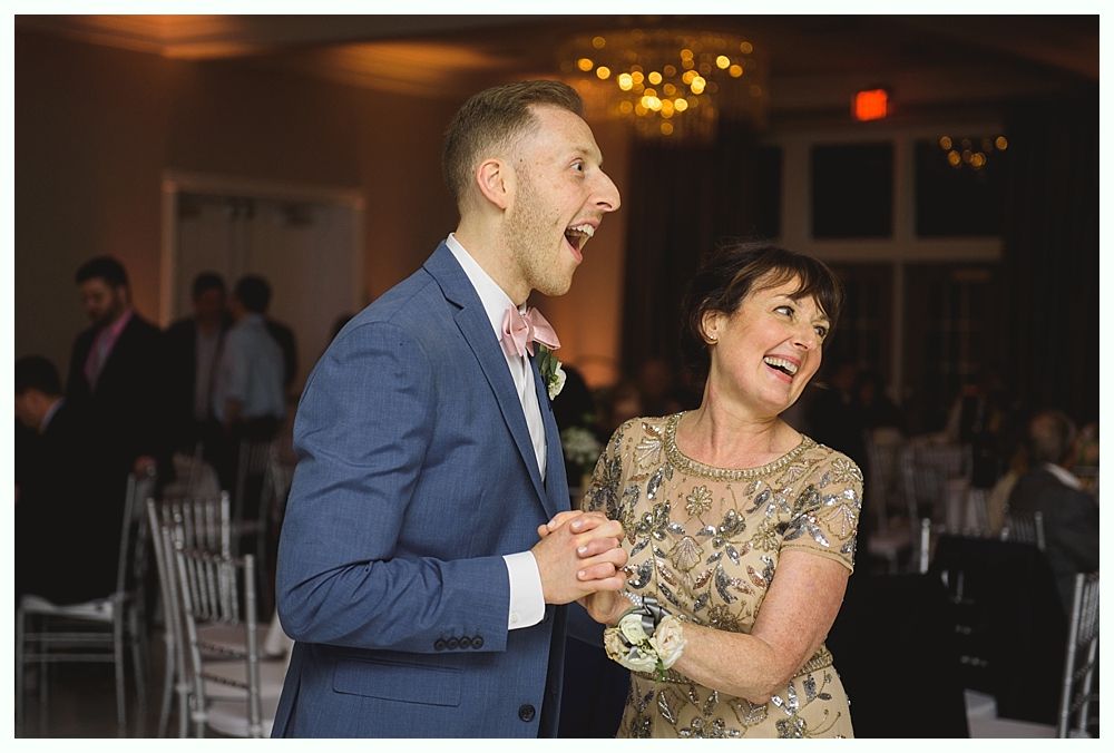 Man and woman laughing, dancing at an event, man in blue suit, woman in embellished dress, indoor setting.