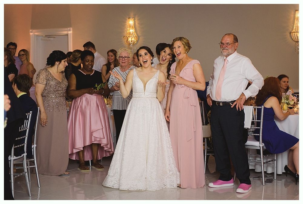 Bride in white gown cheers with wedding party in a reception hall; guests observe.