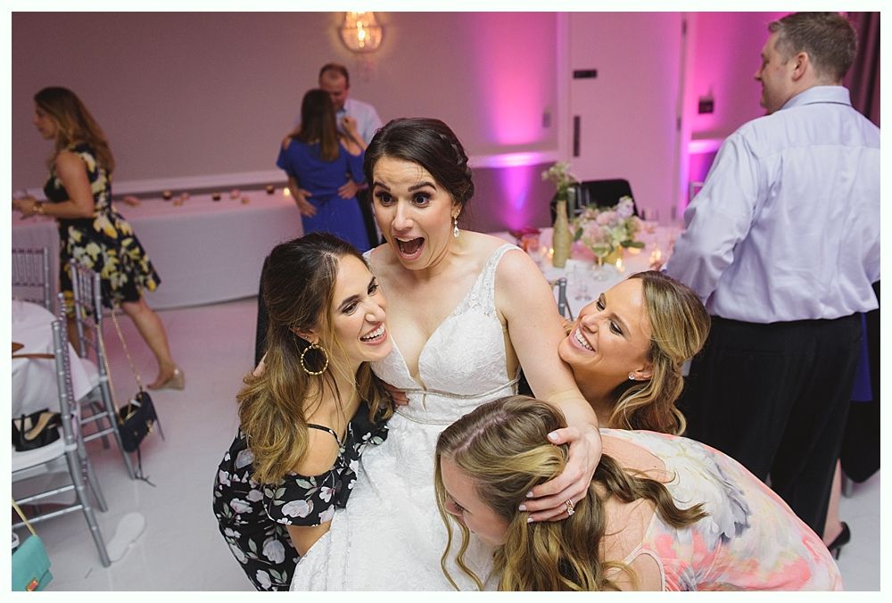 Bride surrounded by laughing women, shocked expression, in a reception hall.