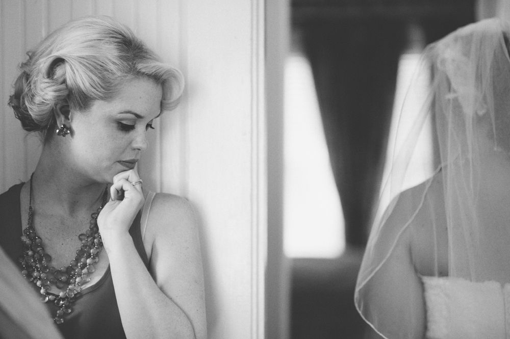 Bride and groom embrace outdoors, groom kisses bride's cheek. Black and white photo with blurred forest background.