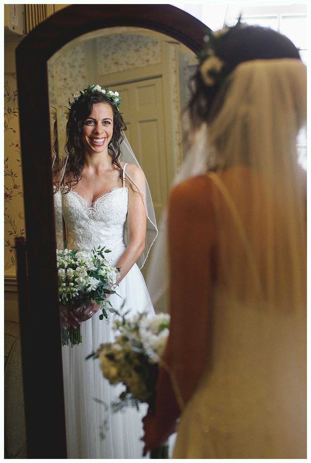 Bride in wedding dress, looking in mirror with bouquet; soft smile.