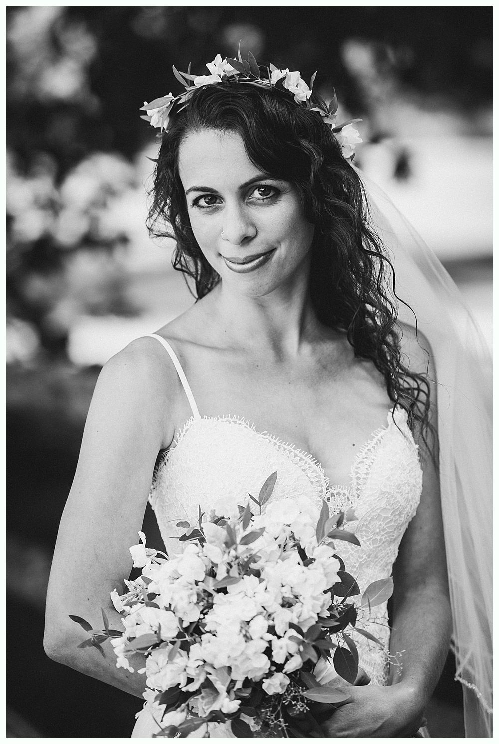 Bride in a beaded wedding dress, holding a bouquet, wearing a floral crown and veil, smiling.