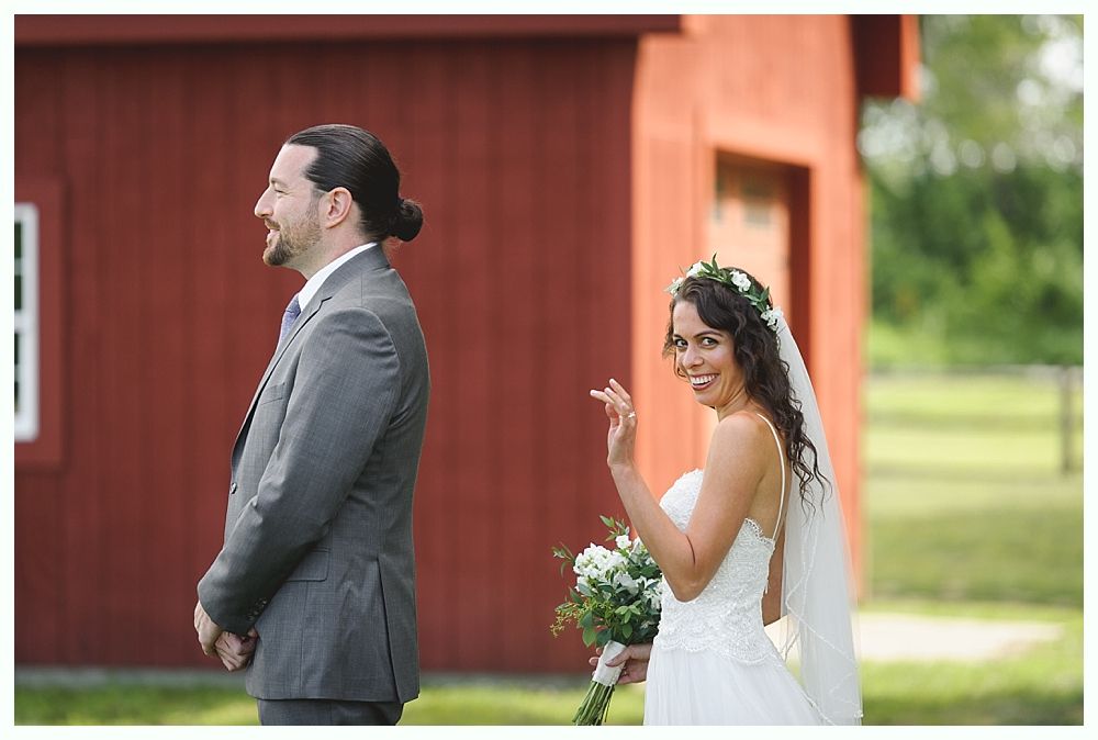 Bride waves at groom before a red barn. She holds a bouquet. He smiles.