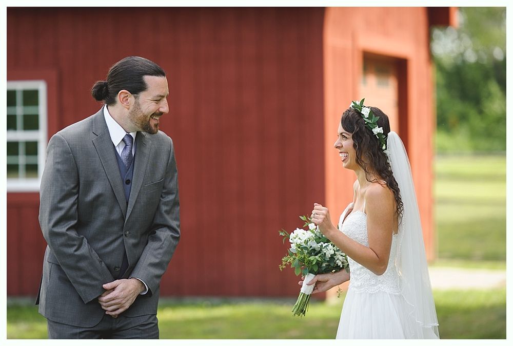 Bride and groom smiling at each other. He wears a gray suit, she has a white dress and veil, a bouquet. Red barn background.