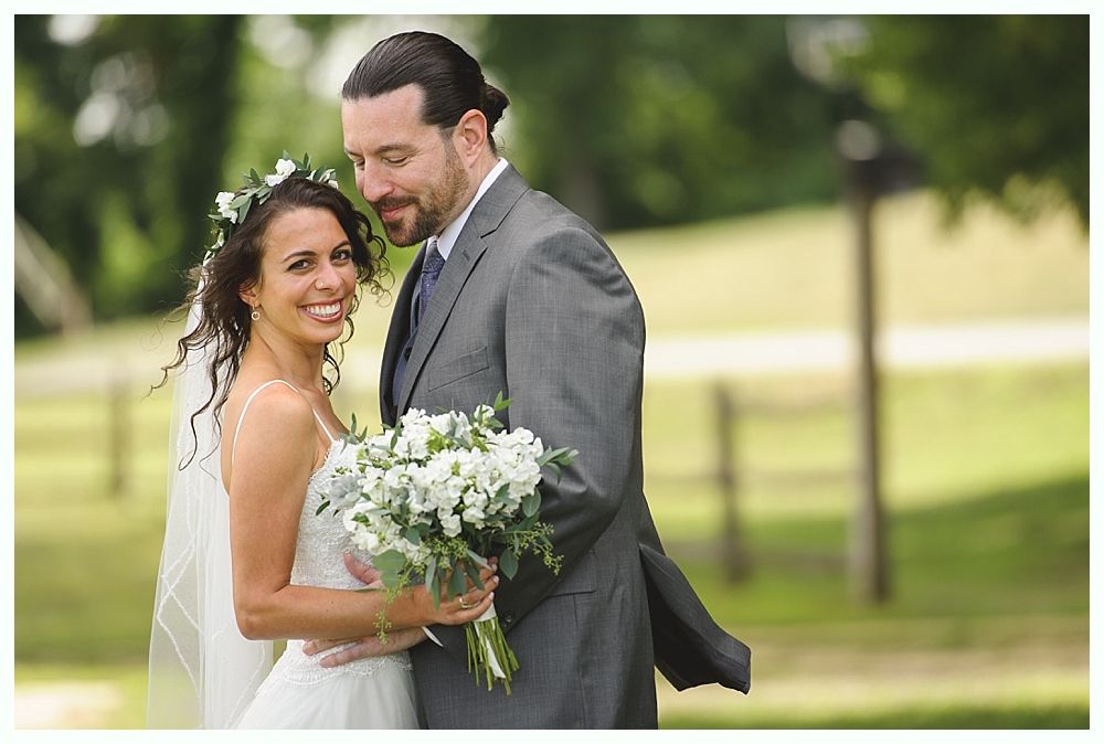 Bride and groom smiling, holding flowers, outdoors. She wears a white dress and veil, he wears a gray suit.
