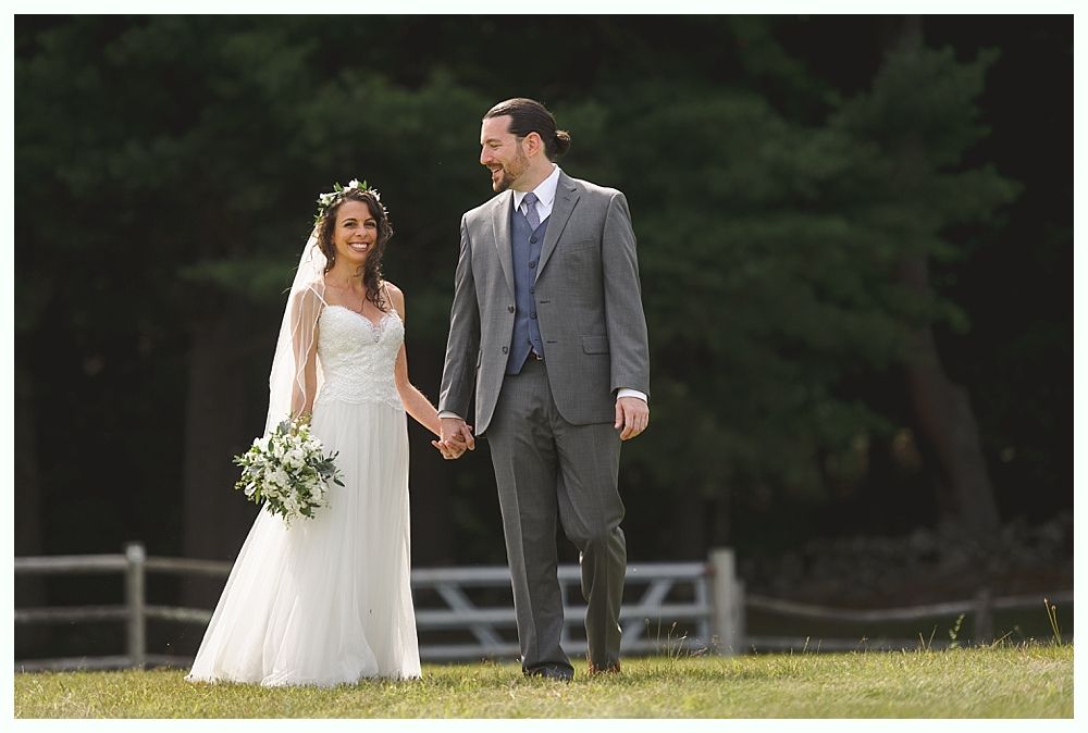 Bride and groom holding hands, smiling, walking on grass. The bride wears a white dress and veil, groom in a grey suit.