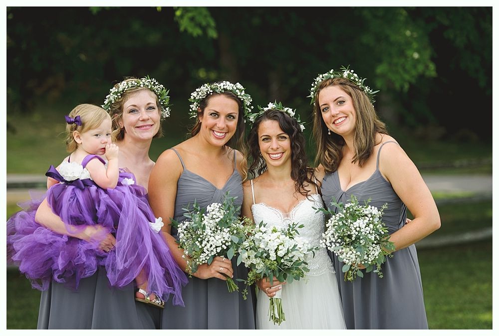 Bride with bridesmaids and flower girl holding bouquets, wearing floral crowns and gray dresses; outdoor setting.