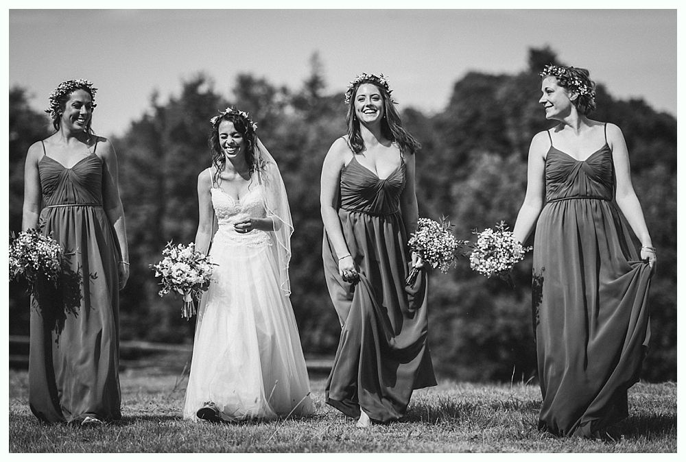Bride with bridesmaids in flowing dresses, walking on grass in front of trees.