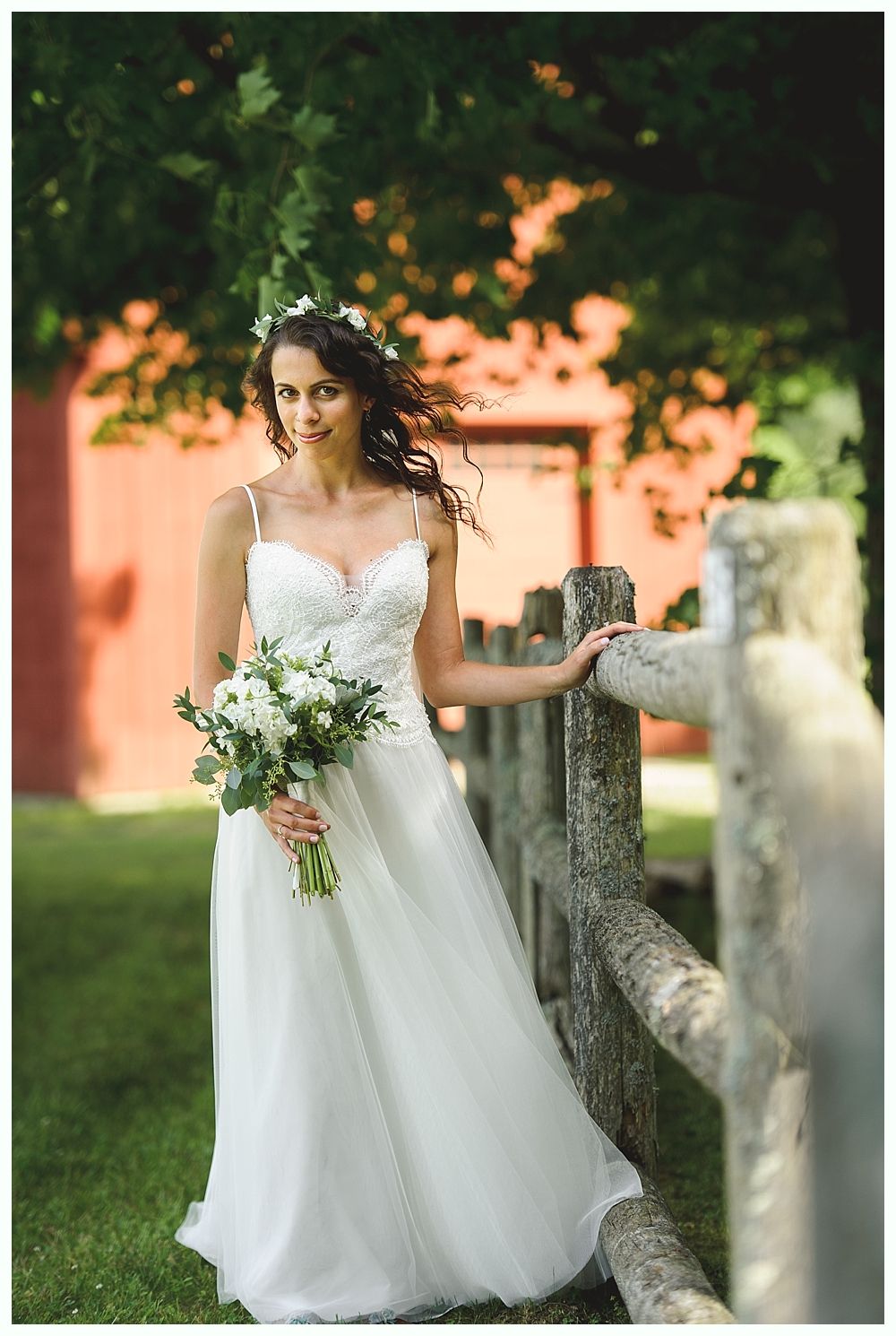 Bride in a white gown and floral headpiece, holding a bouquet, leans on a wooden fence near a red building.