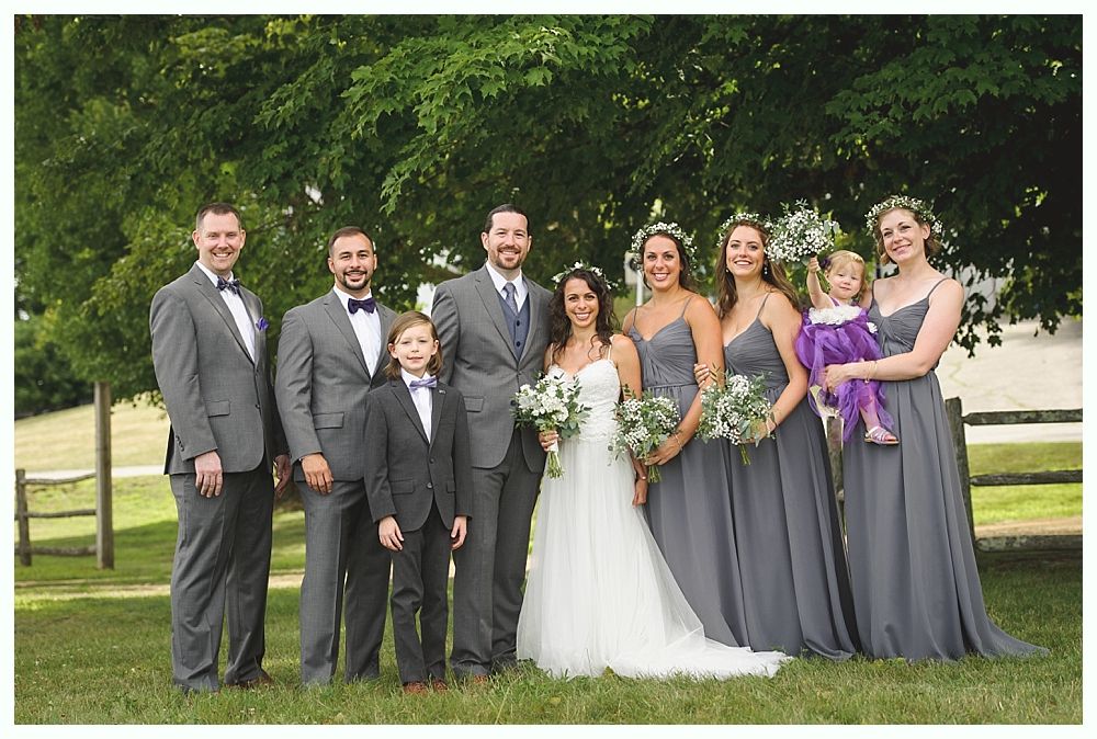 Wedding party posing outdoors; bride in white gown, bridesmaids in gray, groomsmen in gray suits, child in purple.