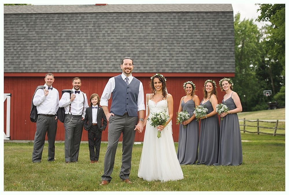 Wedding party poses in front of a red barn. Bride and groom hold hands; bridesmaids wear gray dresses. Groomsmen are in vests.