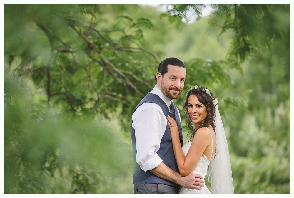 Couple embracing outdoors, bride in wedding dress with veil, groom in vest and tie, surrounded by greenery.