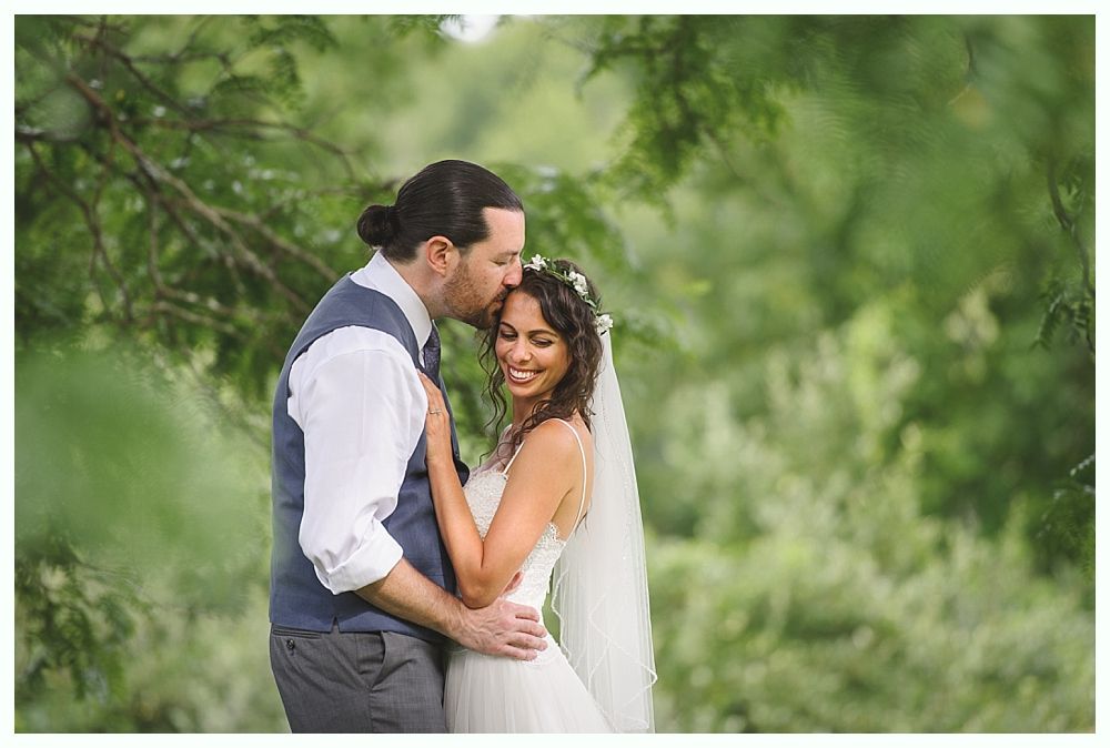 Groom kisses bride's forehead; they embrace, smiling in front of green foliage.