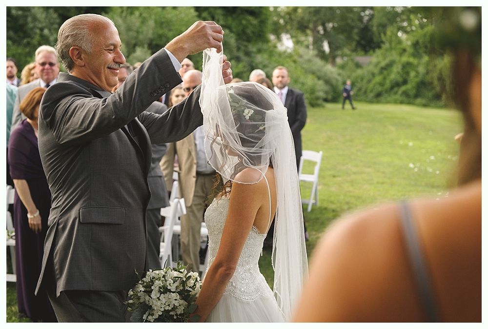 A man adjusts a wedding veil on a bride during an outdoor ceremony. Guests watch from behind.