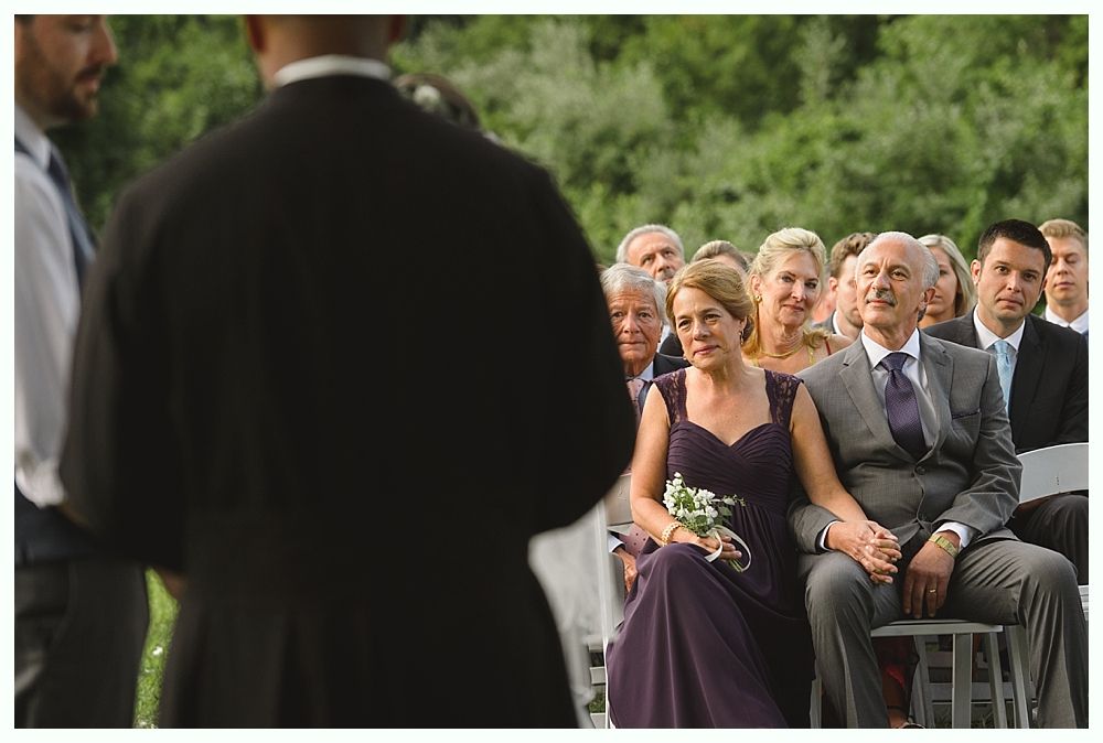 People seated at outdoor wedding ceremony; officiant in front.